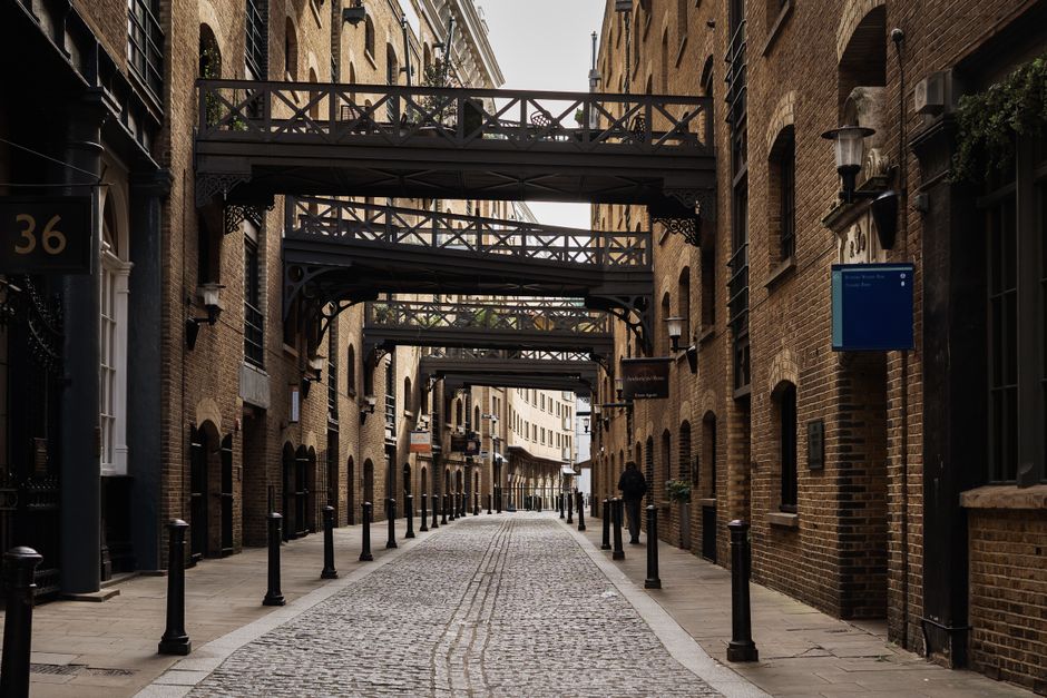 A narrow cobblestone street in an urban area with brick buildings on both sides, featuring multiple black cast iron bridges connecting upper floors of the buildings, used for pedestrian access. The st
