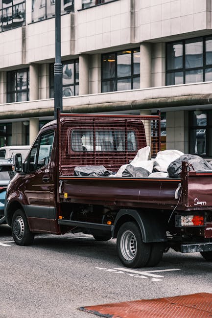 A burgundy flatbed van parked on a street in Rotherhithe, loaded with several large white and grey plastic-wrapped packages, some covered with black plastic bags. The vehicle is positioned near a mode